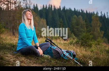 Femme assise sur la pelouse et se reposant pendant la marche avec des bâtons et un sac à dos, regardant avec un sourire détendu et insouciant, respirant de l'air frais.Concept de voyage, randonnée. Banque D'Images