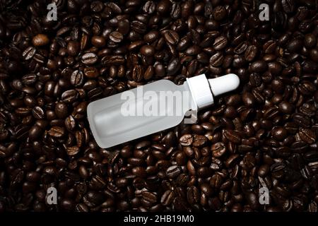 Sérum visage en flacon en verre blanc mat avec compte-gouttes sur fond texturé de grains de café torréfiés. Soin de la peau à base de caféine. Banque D'Images