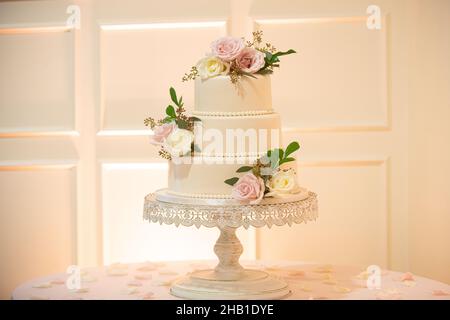 Magnifique gâteau de mariage à trois niveaux avec perles et fleurs roses et blanches sur le plateau à gâteau en dentelle près du mur blanc Banque D'Images