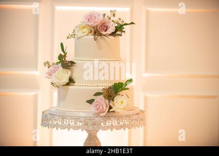 Magnifique gâteau de mariage à trois niveaux avec perles et fleurs roses et blanches sur le plateau à gâteau en dentelle près du mur blanc Banque D'Images