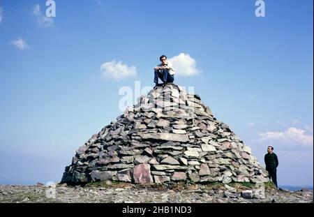 Jeune homme assis sur un cairn de pierre au sommet de Dunkery Beacon, point culminant du parc national d'Exmoor, Somerset, Angleterre 1,705 pi 520 mètres, 1967 Banque D'Images