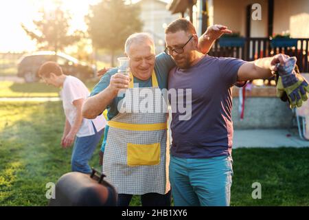 Père en droit avec son fils en droit se serrer, souriant et appréciant le week-end tout en se tenant à côté du grill.Concept de regroupement familial. Banque D'Images
