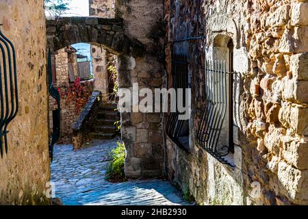 Une ruelle pavée à Le Parage, le quartier médiéval de Les Arcs-sur-Argens, dans le département du Var en France, passe sous une arche de pierre vers une volée de marches ; des barres en fer forgé sécurisent une fenêtre étroite dans le mur à droite, et le feuillage automnal rouge grimpe sur la maçonnerie. Banque D'Images