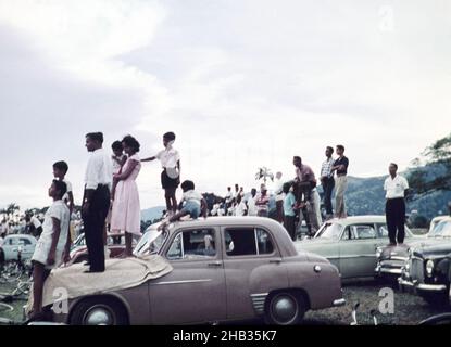 Personnes debout en voiture pour assister à un événement sportif scolaire - match de football annuel entre le Queen Mary's College et le Queen's Royal College - Queen's Park Savannah, Port of Spain, Trinidad c 1962 Banque D'Images