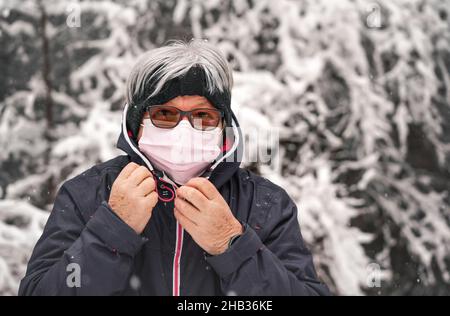 Femme âgée sénior avec cheveux gris portant un masque rose jetable de virus, hiver flou neige couverte arbres fond Banque D'Images