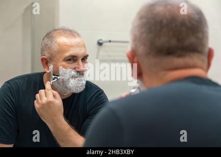 Homme mûr et élégant aux cheveux gris essayant de couper sa barbe dans une salle de bains Banque D'Images