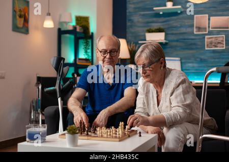 Couple retraité appréciant un jeu de chessboard ensemble assis dans le salon sur le canapé.Mari et femme âgés jouant sur la table comme divertissement à la maison.Vieux avec des béquilles et un cadre de marche Banque D'Images