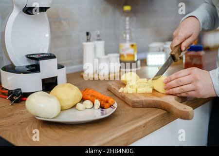 Vue latérale sur les mains d'une femme inconnue avec couteau montez dans la cuisine en coupant des morceaux de pomme de terre pour le soupe végétalienne ou végétarienne cuisant un repas sain Banque D'Images