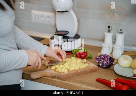 Vue latérale sur les mains d'une femme inconnue avec couteau montez dans la cuisine en coupant des morceaux de pomme de terre pour le soupe végétalienne ou végétarienne cuisant un repas sain Banque D'Images