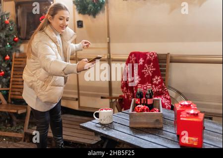 Moscou 11.29.2021. Jeune femme candide tenant dans les mains smartphone et prenant photo pour blog d'une table avec des boissons sur bonne année et victoire de noël Banque D'Images