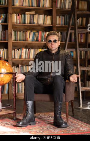 Homme dans une veste en cuir hipster et lunettes de soleil stylées se trouve dans un fauteuil rétro avec des mains ouvertes dans Loft Studio .Arrière-plan des étagères de livres.Gros plan .Photo de haute qualité Banque D'Images