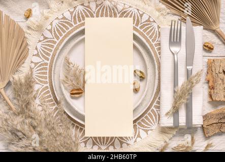 Table de mariage avec carte vierge verticale sur l'assiette sur platemat bohème vue de dessus.Maquette de carte de mariage boho.Modèle beige tropical avec plan séché Banque D'Images