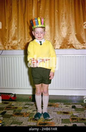 Jeune garçon portant un pull jaune et un chapeau indien rouge jouant avec un jouet de roquette spatiale rouge le matin de Noël, culture britannique 1967 Banque D'Images