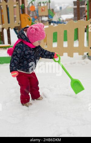 Une petite fille de 2 ans de race blanche marche en hiver sur l'aire de jeux.Un enfant creuse la neige avec une pelle Banque D'Images