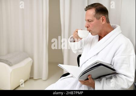 Beau imposant mûr 50 ans homme européen habillé en éponge blanc peignoir, boire un thé à base de plantes saines et lire le magazine assis sur l'armcha Banque D'Images