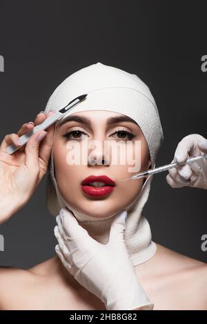 chirurgiens de plastique tenant le couteau chirurgical et la seringue près de la femme dans un bandage élastique sur la tête isolée sur gris foncé Banque D'Images