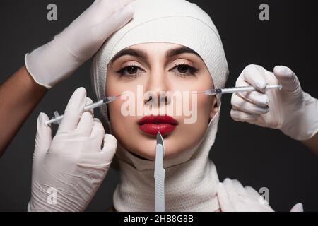 chirurgiens de plastique tenant le couteau chirurgical et les seringues près du patient dans un bandage élastique sur la tête isolé sur le gris foncé Banque D'Images