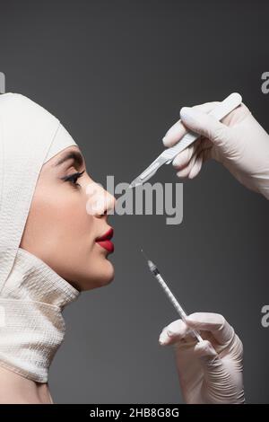 vue latérale du patient dans un bandage élastique près des chirurgiens en plastique tenant un couteau chirurgical et une seringue isolés sur le gris foncé Banque D'Images