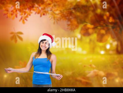 Image composite d'une femme caucasienne portant un chapeau de père noël avec du ruban de mesure autour de sa taille contre le parc Banque D'Images