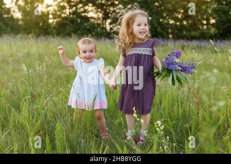 Mignons, petites filles en robes tiennent dans les mains bouquet de fleurs lupin et sauter.Enfants souriants et amusants sur le terrain.Les enfants jouent à l'extérieur.Concept de hap Banque D'Images
