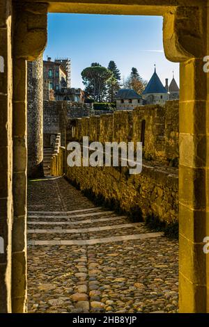 Une ouverture de fenêtre en pierre dans le mur d'enceinte de la Cité de Carcassonne, département de l'Aude, en France, encadre une vue sur un chemin pavé entre les murs ; au fond, un bâtiment sous échafaudage et une tour au toit pointu. Banque D'Images