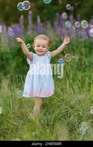 Jolie petite fille en robe bleue essayer de attraper des bulles de savon.Bébé souriant s'amusant parmi les fleurs lupin.Jeux pour enfants en extérieur.Concept de chi heureux Banque D'Images