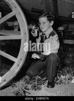 Jeune garçon dans la tenue de cow-boy agenouillé à côté de roue en bois sur stagecoach regardant le canon d'un fusil de jouet.Vers 1955 Banque D'Images