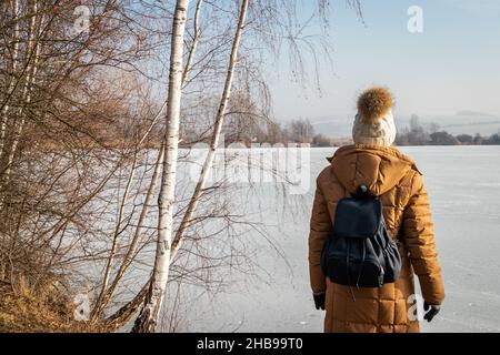 Une femme portant un manteau d'hiver et un chapeau tricoté est debout sur un lac gelé et un bouleau.Randonnée dans la nature par temps froid et vue imprenable Banque D'Images