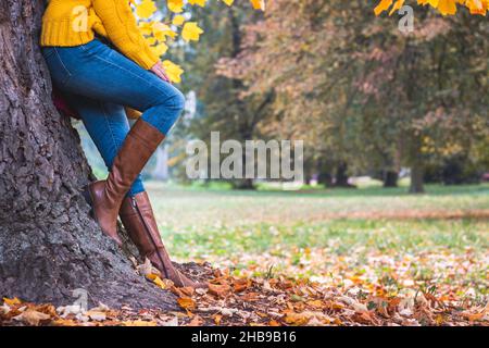 La femme de mode est penchée sur l'arbre au parc d'automne.Jambes de femme portant un Jean et une botte en cuir.Vêtements tendance et décontractés Banque D'Images