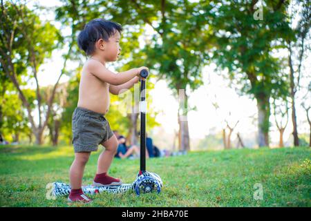 Adorable garçon jouant le scooter sur l'herbe verte dans l'éréation du parc cet été Banque D'Images