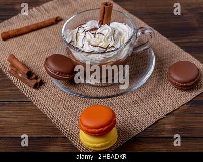 Une tasse de verre avec du chocolat chaud et de la crème sur une table avec quelques macarons colorés Banque D'Images