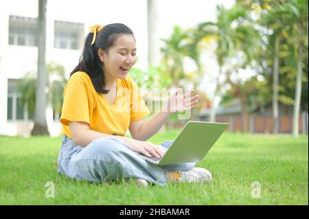 Jeune femme assise sur la pelouse discutant avec des amis s'amusant sur la pelouse, portant une chemise jaune et un Jean, visage souriant, ordinateur portable sur les jambes, travail de fond Banque D'Images