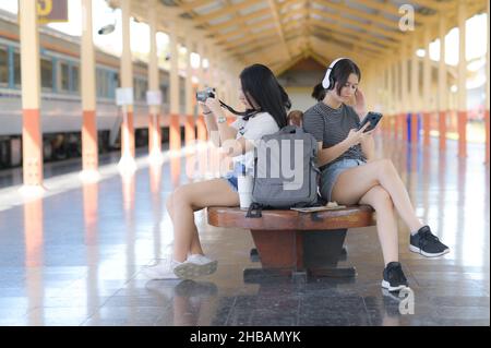 Deux adolescentes attendant un train sur une plate-forme pour un voyage, une femme asiatique dans une chemise blanche prenant un appareil photo pour prendre une photo, une femme européenne Banque D'Images