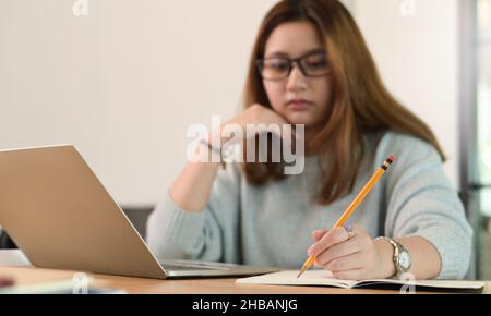 Une adolescente portant des lunettes utilise un crayon pour écrire sur un bloc-notes avec un ordinateur portable sur le bureau, une étudiante d'université prenant des notes en ligne d'un tour Banque D'Images