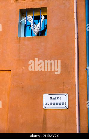 Mur de maison avec une grille de fenêtre derrière laquelle se dresse la sculpture d'un coq.Un panneau avec le nom de la rue: 'Traverse des Troubadours' (carrefour de Minsrel).Bormes-les-Mimosas, France Banque D'Images