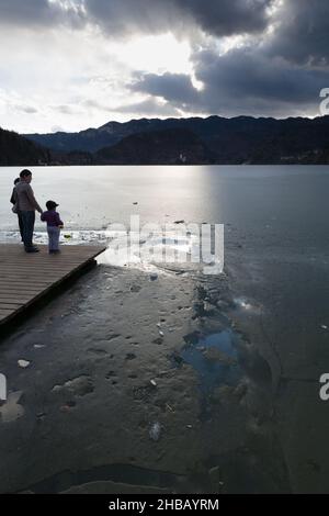 Une femme et ses deux enfants se tenant sur une jetée qui fait face à Frozen Lake Bled jusqu'à l'église de pèlerinage de l'Assomption de Marie, Sloven Banque D'Images