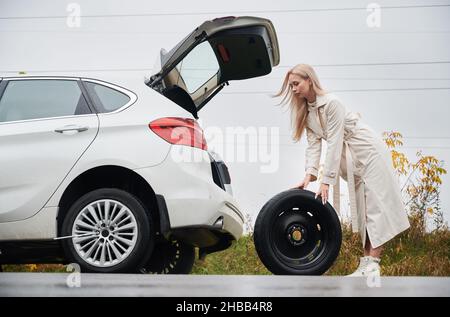 Vue latérale d'une femme blonde se tenant à côté de la voiture avec le coffre ouvert, tenant la roue de secours dans ses mains pour remplacer la roue arrière de la voiture endommagée et dégonflée. Banque D'Images