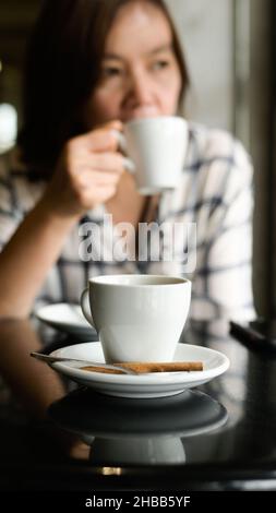 Gros plan Une tasse de café blanc est placée sur la table, une jeune femme s'assoit à boire du café en arrière-plan. Banque D'Images