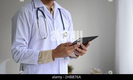 Coupe courte d'UN professionnel de la santé dans une blouse de laboratoire avec un stéthoscope tenant un comprimé à la main, vue latérale. Banque D'Images