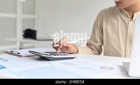 Un jeune homme avec un stylo à la main utilise une calculatrice, des données graphiques et un ordinateur portable sur le bureau. Banque D'Images