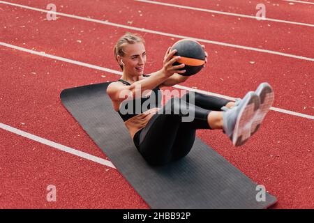 Jeune femme de coupe perfectionne son corps tout en faisant de l'exercice avec Medicine ball sur le tapis de piste de stade avec revêtement rouge.Un mode de vie sain Banque D'Images