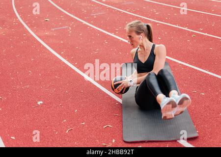 Jeune femme de coupe perfectionne son corps tout en faisant de l'exercice avec Medicine ball sur le tapis de piste de stade avec revêtement rouge.Un mode de vie sain Banque D'Images