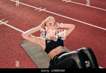 La jeune femme en forme de femme perfectionne son corps en faisant de l'exercice de torsion pour les muscles abdominaux sur le tapis de piste du stade avec revêtement rouge par temps ensoleillé.Santé l Banque D'Images