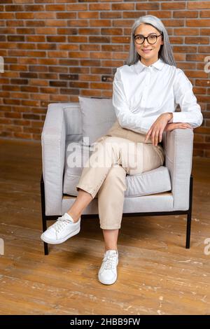 Vue en longueur de la femme âgée moderne, positive et saine, assise dans le fauteuil du salon moderne et regardant la caméra.Charmante femme âgée rêvant.Concept de mode de vie sain Banque D'Images