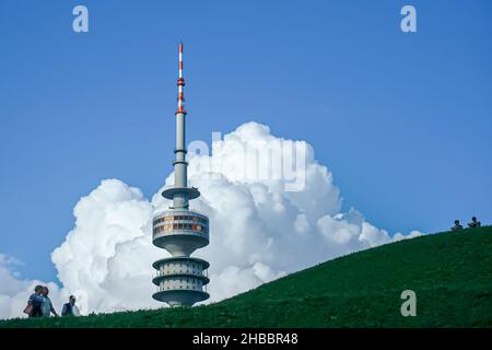 Un couple et un homme marchent sur une colline près de la Tour Olympique de Munich.Deux jeunes hommes s'assoient en haut à droite.Grands nuages dans le ciel bleu. Banque D'Images