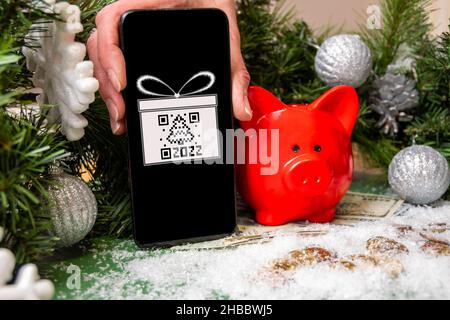 Une main tenant un smartphone avec un symbole cadeau et un code QR en forme de sapin, une banque de porc rouge sur des pièces d'or recouvertes de neige et de Christma Banque D'Images