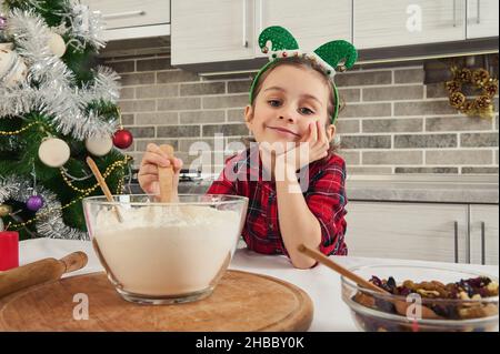 Enfant mignon, belle fille sourit, regardant l'appareil photo de rêve, mélangeant des ingrédients secs dans un bol en verre.Adorable petit chef prépare des biscuits Banque D'Images