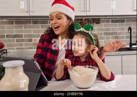 Portrait d'adorable belle jolie petite fille gaie mignonne et sa charmante maman en chapeau de Santa mélangeant les ingrédients dans un bol avec des précordiaux de farine Banque D'Images