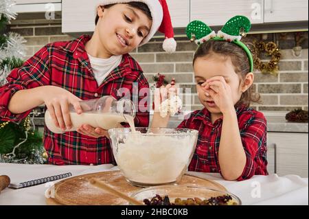 Beau préadolescent de race blanche vêtu d'une chemise à carreaux rouge et vert et d'un chapeau de père Noël verse du lait dans un bol, cuisant avec sa petite sœur Banque D'Images