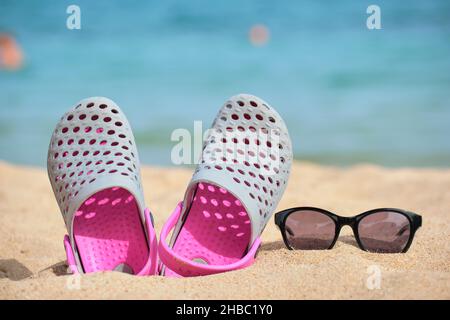 Gros plan sur les chaussures Clogs et les lunettes de soleil noires protectrices sur la plage de sable au bord de la mer tropicale par temps chaud et ensoleillé. Concept de vacances d'été. Banque D'Images